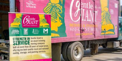 Purple moving truck with a logo of giant yellow feet and the words 'Gentle Giant Moving Company' written on it. Beside the truck is a sign with information about the Gentle Giant company.