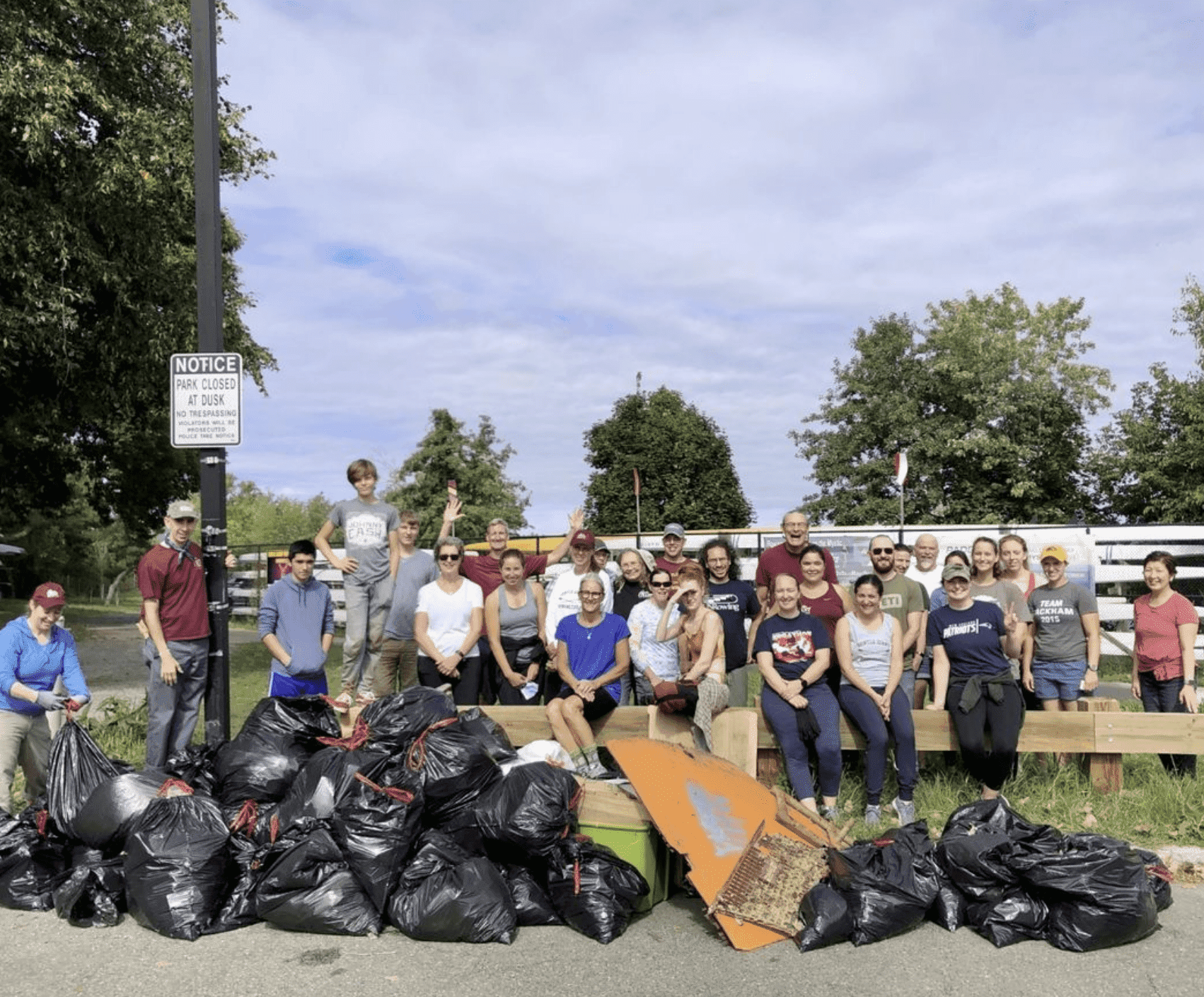 A group of people are pictured standing together near filled black bin bags. The black bin bags are filled with trash they have collected from the Charles River on a river clean event.