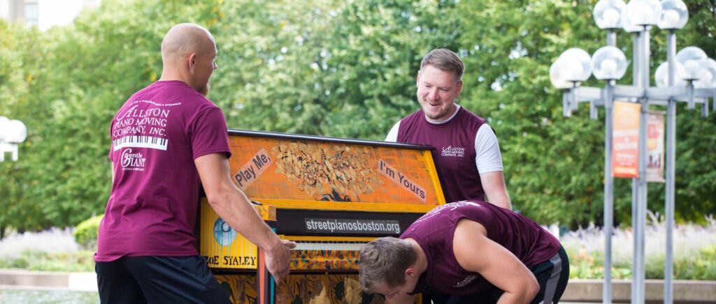 Three movers are rolling an orange piano down the street. Each mover is dressed in a maroon shirt and black pants.