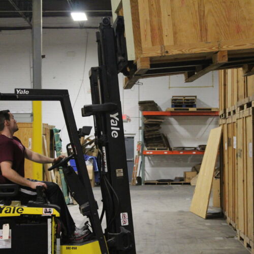 A man on a forklift is lifting a wooden vault on top of another wooden vault in a storage warehouse.