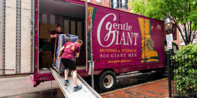 Two movers pushing a large piece of furniture down the ramp at the side of a purple moving truck.