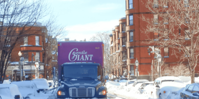 A Gentle Giant moving truck is park on a snow-covered street in Boston. There are brownstone buildings lining the street.