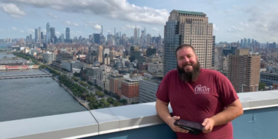 A man is dressed in a maroon shirt and black shorts. He is standing on a balcony. In the background is the New York City skyline.