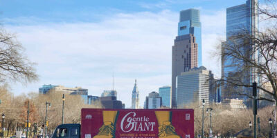 A Gentle Giant purple moving truck is parked horizontally on a walking path. Skyscrapers are visible in the background.