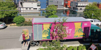 A Gentle Giant mover is walking up a ramp connected to a Gentle Giant moving truck. He his carrying two boxes. The purple moving truck is parked in front of a house. In the background, behind the moving truck, there are houses and a lake with boats on it.
