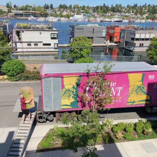 A Gentle Giant mover is walking up a ramp connected to a Gentle Giant moving truck. He his carrying two boxes. The purple moving truck is parked in front of a house. In the background, behind the moving truck, there are houses and a lake with boats on it.