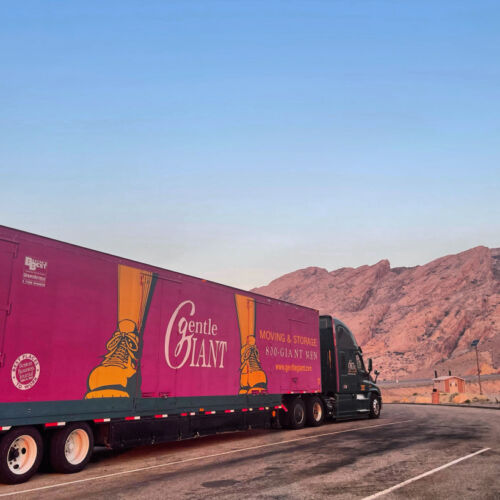 A Gentle Giant Moving & Storage semi-truck with a vibrant magenta and green trailer is parked at a scenic rest stop against a backdrop of rugged, reddish-brown mountains and a clear blue sky. The truck features the company's logo and accolades, including "Best of Boston" and "BBB" ratings. The scene is illuminated by warm, golden-hour lighting, highlighting the truck and the surrounding desert landscape.